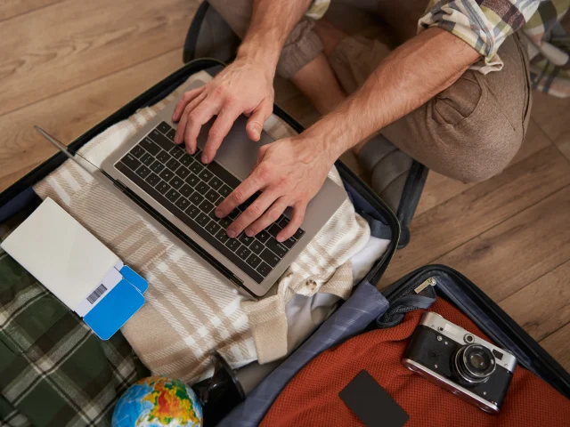 close-up-shot-male-hands-tourist-using-laptop-sitting-floor-with-opened-suitcase-packed